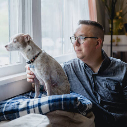 Portrait of an autistic man with his dog indoors. They are sitting on their couch in the living room of his home. Shot using natural window light.